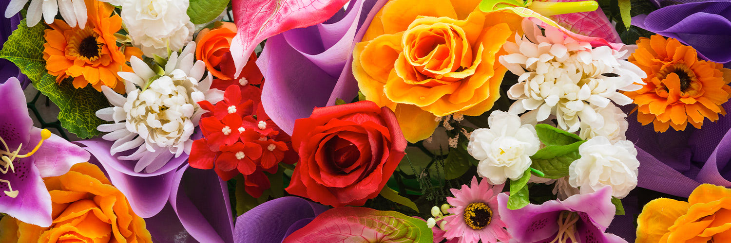 A vibrant arrangement of various colorful flowers, with roses, daisies, and lilies, surrounded by green leaves and purple ribbons in a close-up, detailed view.