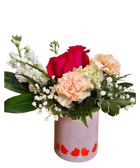 a pink cylinder decorated with red heart lollipops is filled with pink carnations, pink alstroemeria, white daisies, greenery and a heart pick