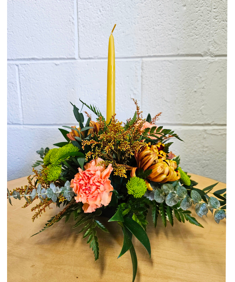 a single taper candle nestled in a Thanksgiving arrangement of orange carnations, fall chrysanthemums, green button mums and fall foliage and accents